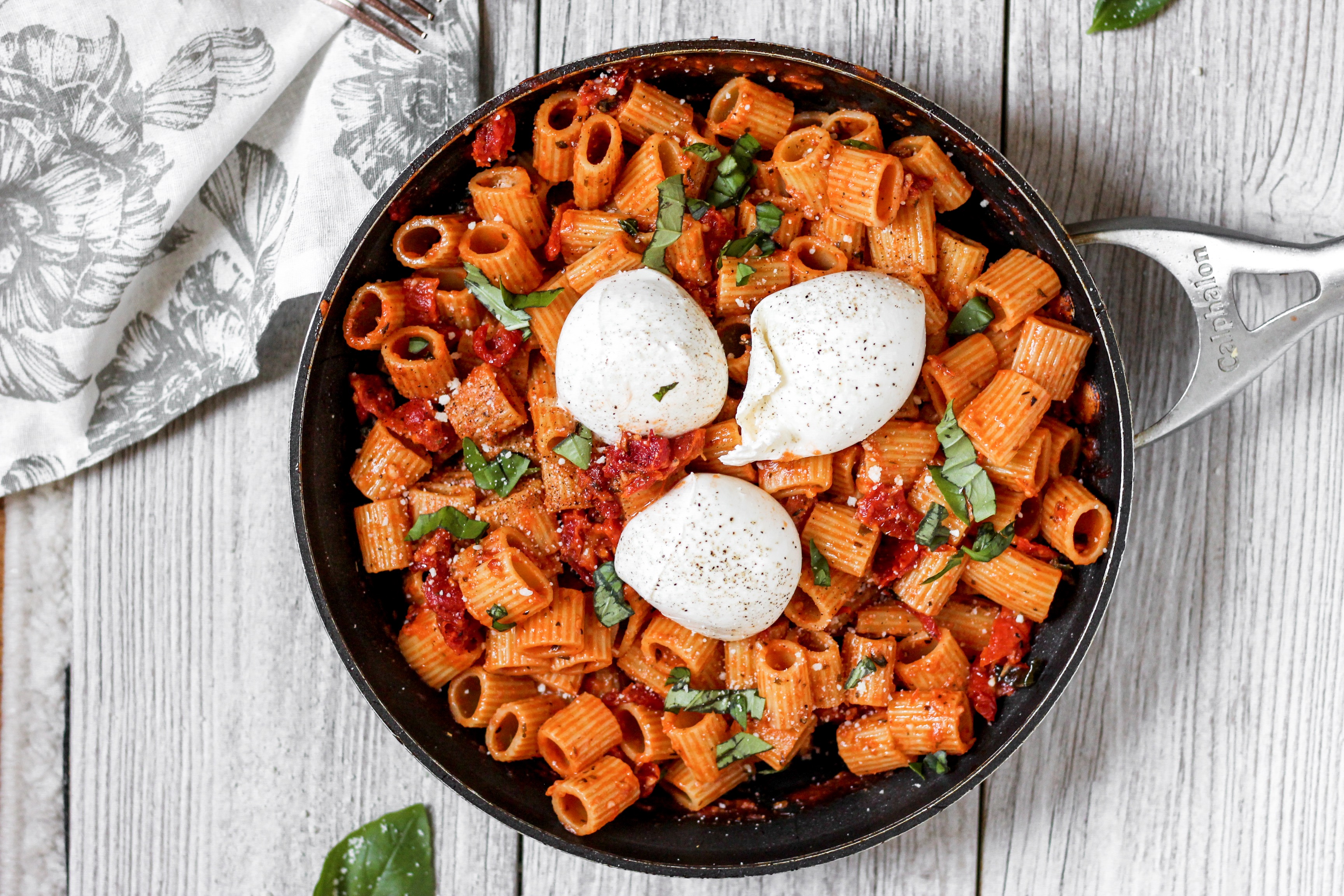 overhead shot of the pasta in a pan on a pale wood surface with a floral pattern linen and a leaf of basil.