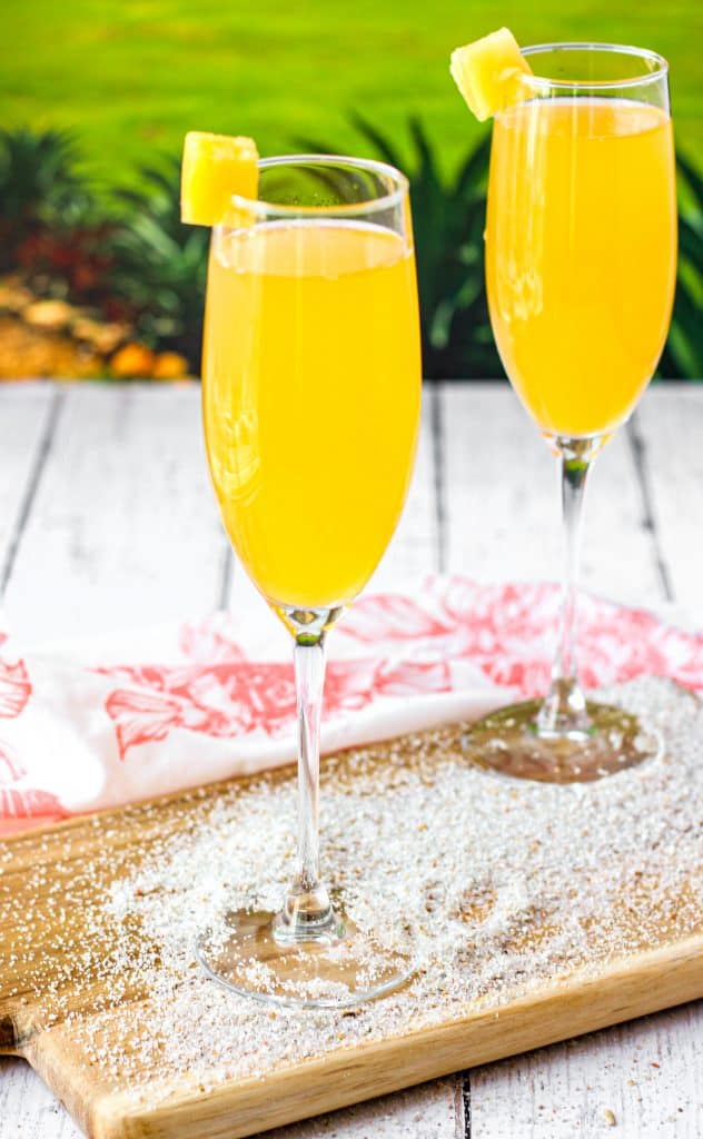 Two peach mimosas on a wood board with sand and a green background.