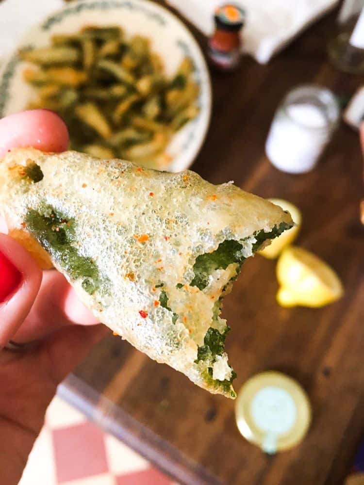 a hand holding the fried sage fritter over a wood board. In the background, slightly blurred images of cut lemons and a bowl of the fried sage.