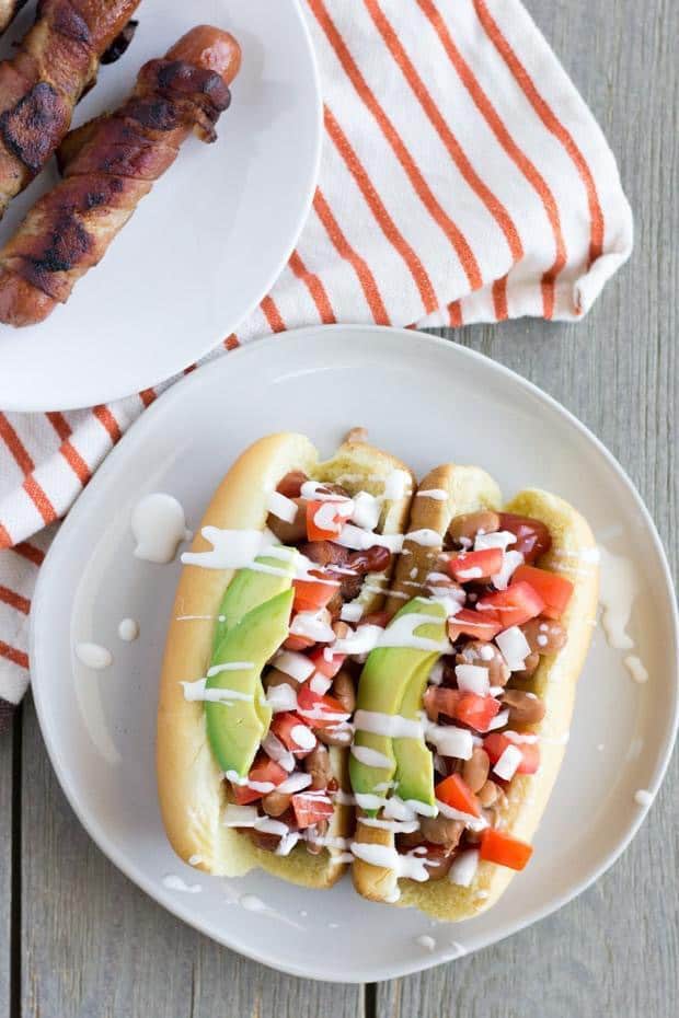 Overhead shot of two of the hot dogs on an off-white plate. Hot dogs are topped with tomatoes, onions, avocado and a white sauce. To the back left, a plate of the grilled hot dogs and an orange stripped napkin. 