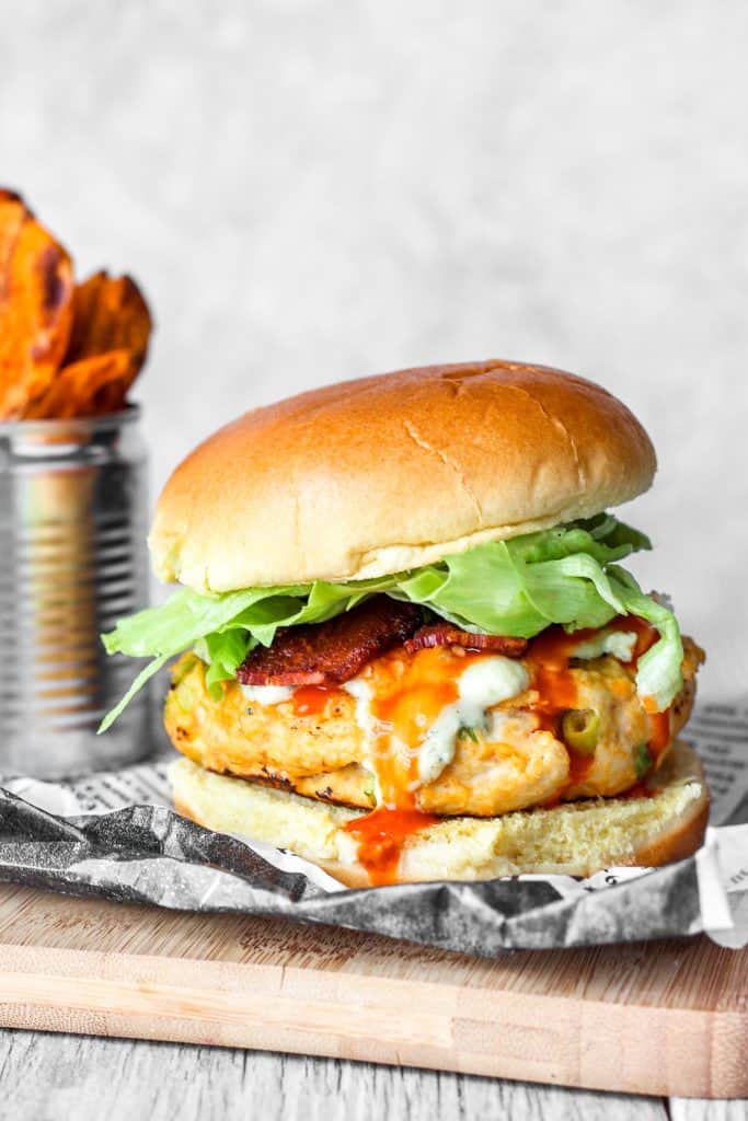 buffalo chicken burger on wood board with sweet potato fries. Crumpled newspaper wax paper under the burger.