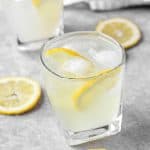 Two rocks glasses with the limoncello cocktail on a grey cement board. Lemons scattered around. In the background a silver jigger and a grey floral print napkin.