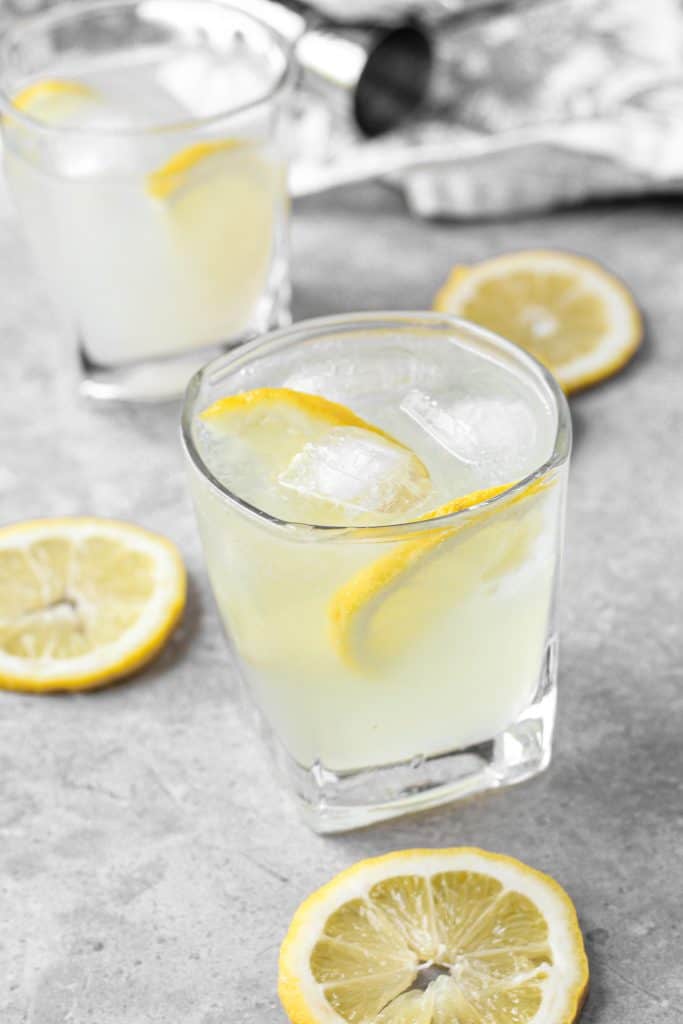 Two rocks glasses with the limoncello cocktail on a grey cement board. Lemons scattered around. In the background a silver jigger and a grey floral print napkin.