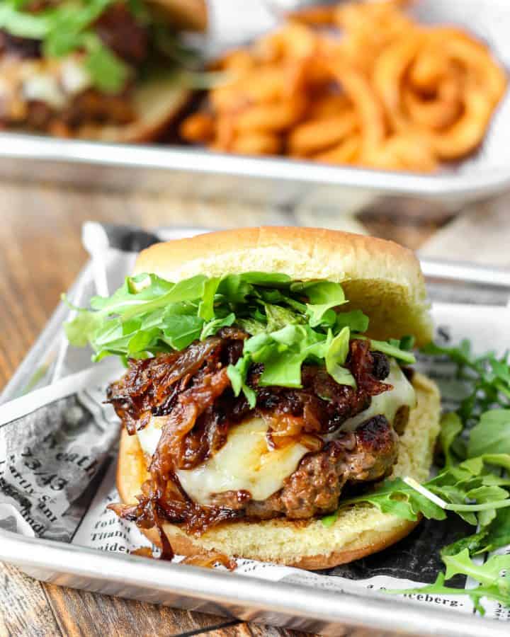 In the foreground one of the french onions burgers topped abundantly with arugula on a metal serving tray with a newspaper wax paper and curly fries. In the background a glass of water, and another burger on the silver tray. All of this sits on a wood patterned board.