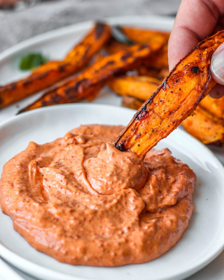 A hand dipping a sweet potato fry into the spicy harissa mayo on a white circular plate.