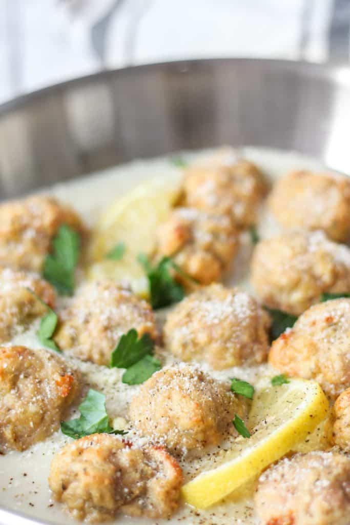 Chicken piccata meatballs in a stainless steel pan with a lemon wedge.