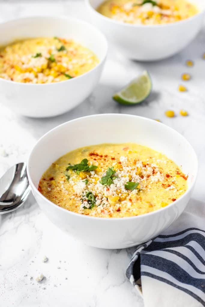 three bowls of the Mexican corn chowder with a spoon and a blue striped linen napkin on a white marble surface.