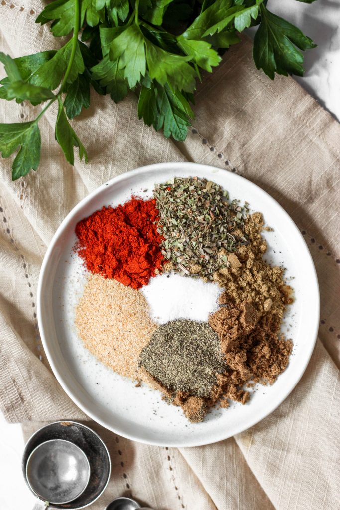 overhead shot of the white round plate with the unmixed spices. Sitting on a beige linen napkin and a bunch of parsley and measuring spoons.