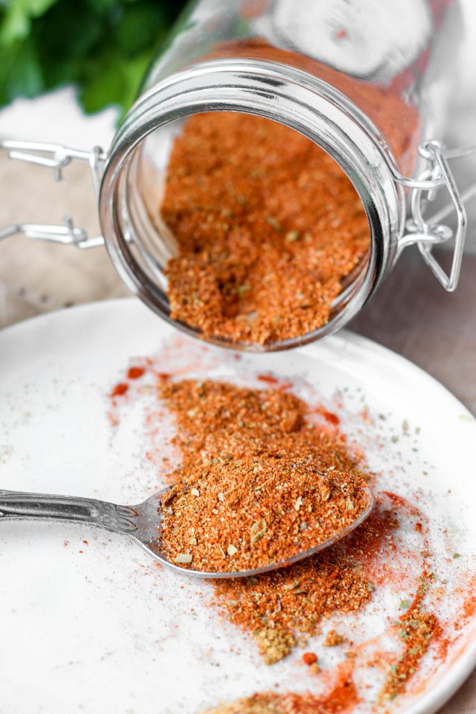 a spoon on a white round plate with the sazon, a jar of sazon on it's side in the back.