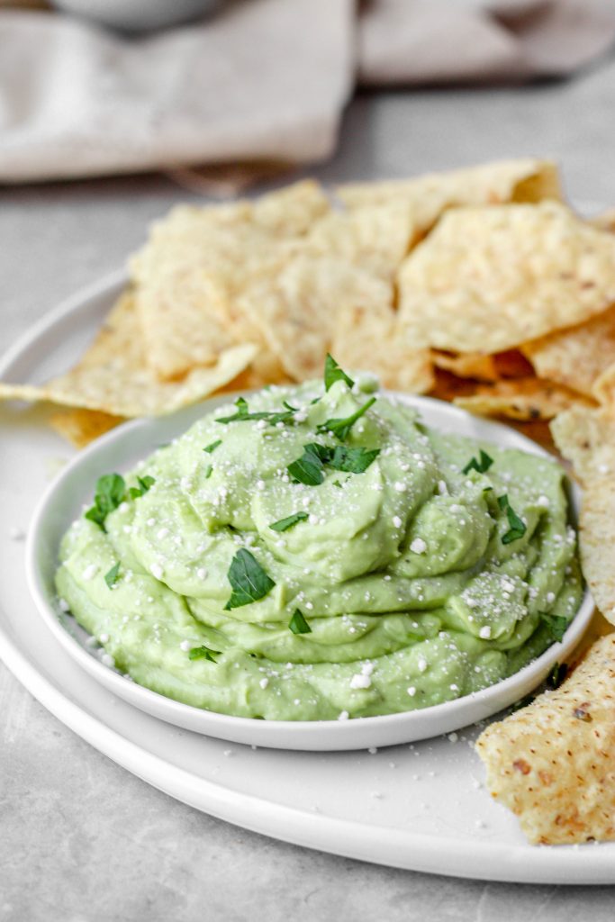 A small plate of the whipped avocado on a round plate with chips. On a cement pattern surface.