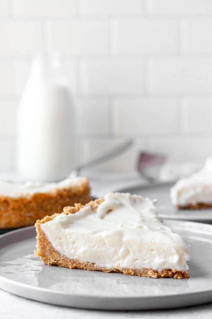 a slice of the cheesecake on a grey plate. In the back the whole cheesecake, another slice on a plate, and a glass of milk. 