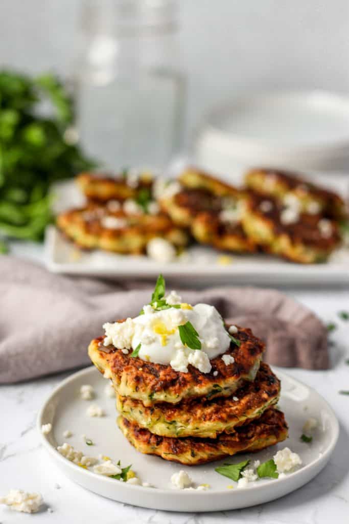 stack of 3 kolokithokeftedes on a white round plate. A plate of fritters in the back.