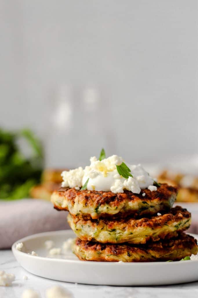 close shot of the 3 fritters on a round plate topped with Greek yogurt and feta and parsley and a little lemon zest.