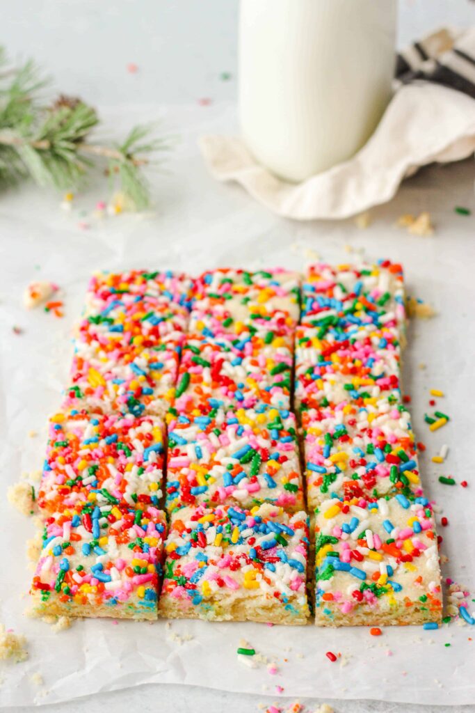 45 degree angle shot of a large rectangle of the sliced cookies on parchment paper, sprinkles everywhere, milk and evergreen in the background.