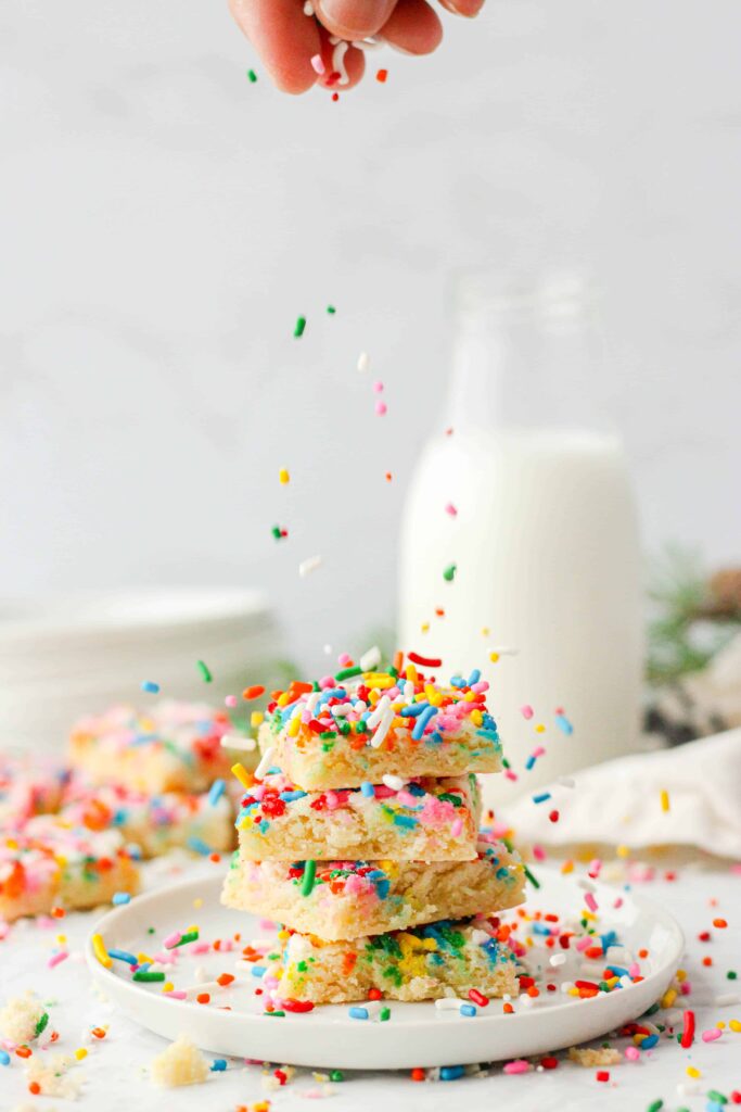 shot of 4 stacked cookies with a hand sprinkling sprinkles. Milk, plates, evergreen and a beige linen in the back.