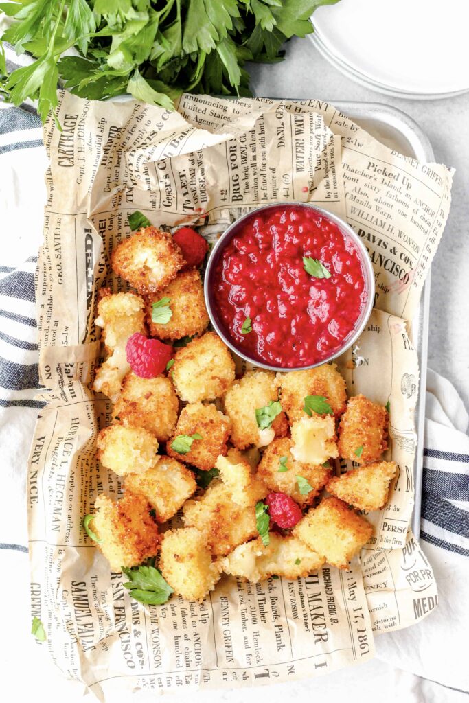 overhead shot of the fried brie bites in a platter with the raspberry sauce, on beige newsprint paper, and a blue striped linen napkin beneath,