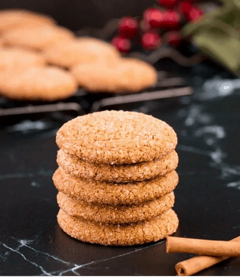 a stack of 5 gingersnaps on a black marble surface. More cookies in the background, 2 cinnamon sticks in the foreground.