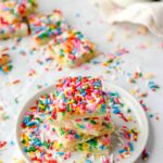 shot taken at 45 degree angle of a stack of 4 square sugar cookies on a round white plate, sprinkles everywhere, some cookies in the background.
