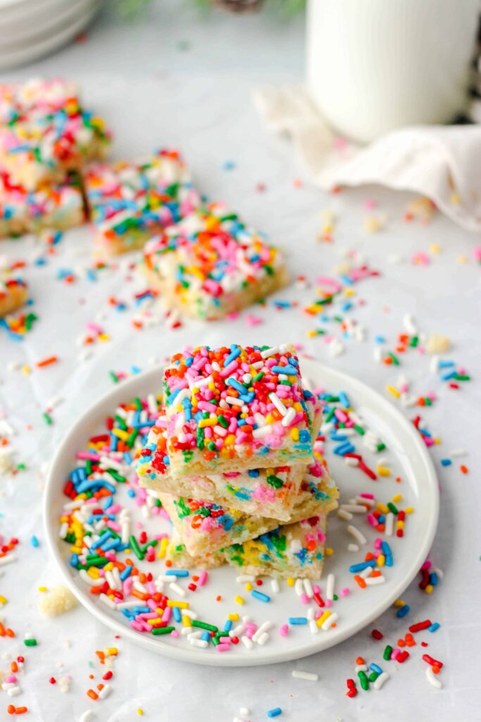 shot taken at 45 degree angle of a stack of  4 square sugar cookies on a round white plate, sprinkles everywhere, some cookies in the background.