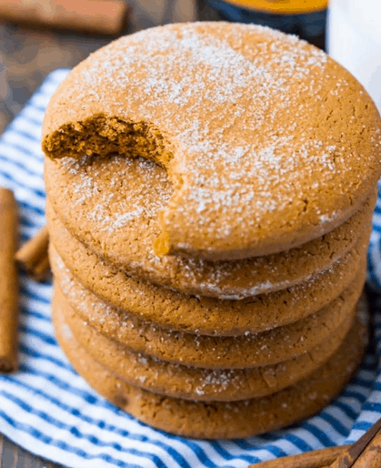 stack of 5 ginger cookies with the top cookie having a bite taken out. Sitting on a blue and white striped linen and cinnamon sticks.