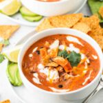 two white bowls of the creamy chicken tortilla soup with yellow corn chips, slices of avocado, limes, and a spoon.