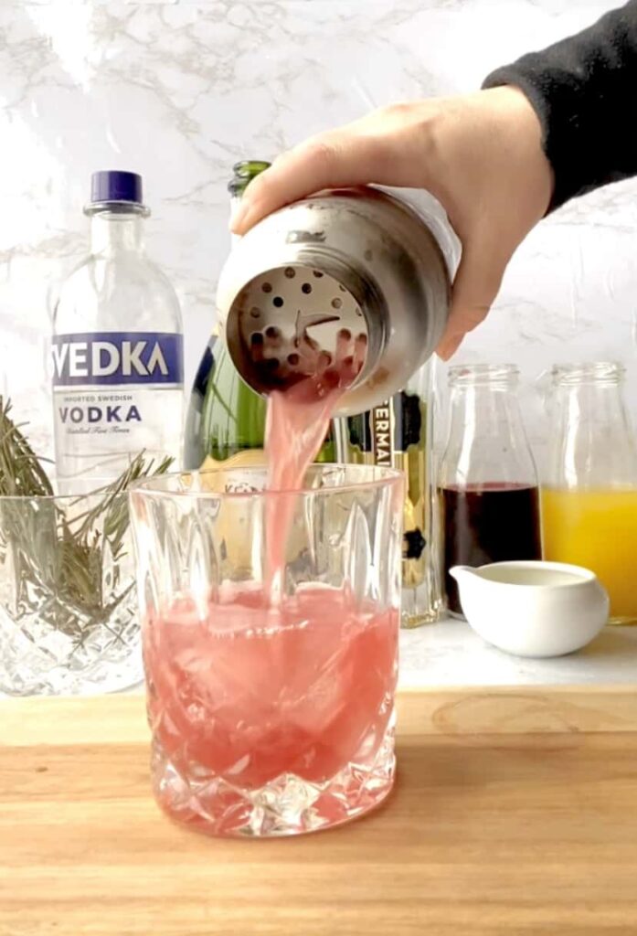 pouring the cocktail into a glass, in the background a bottle of vodka, champagne, elderflower liquor, pomegranate juice, and orange juice, rosemary simple syrup in a small white dish, and a glass of rosemary springs.