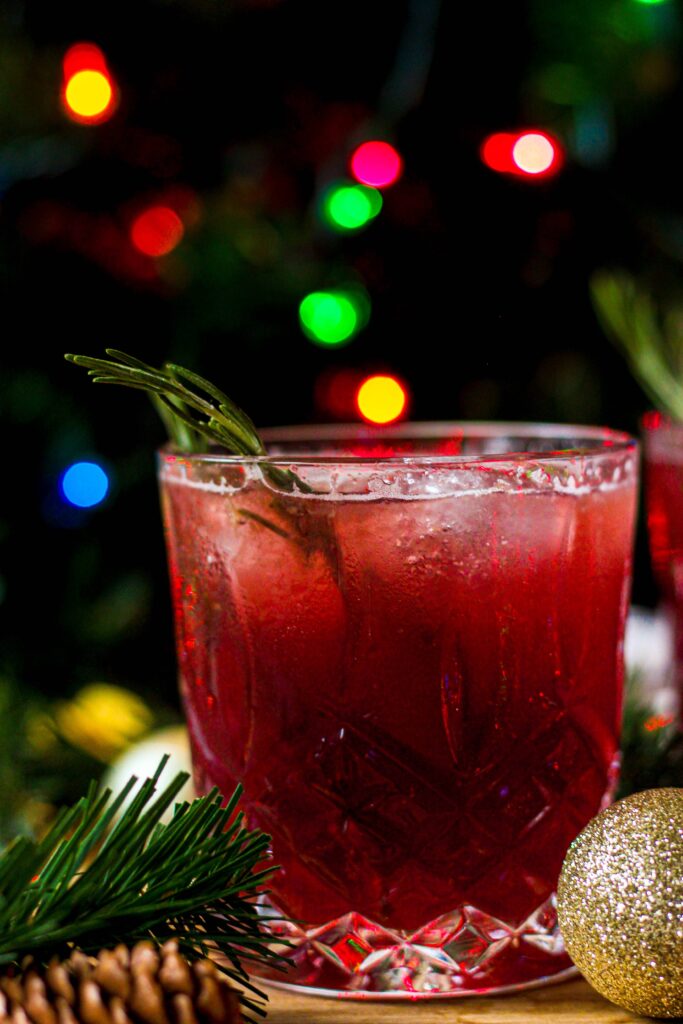 similar to the first photo, one elderflower sparkling cocktail with a christmas theme, evergreen in the back with blurred colored lights and in the front a sparkly gold ornament and more evergreen.