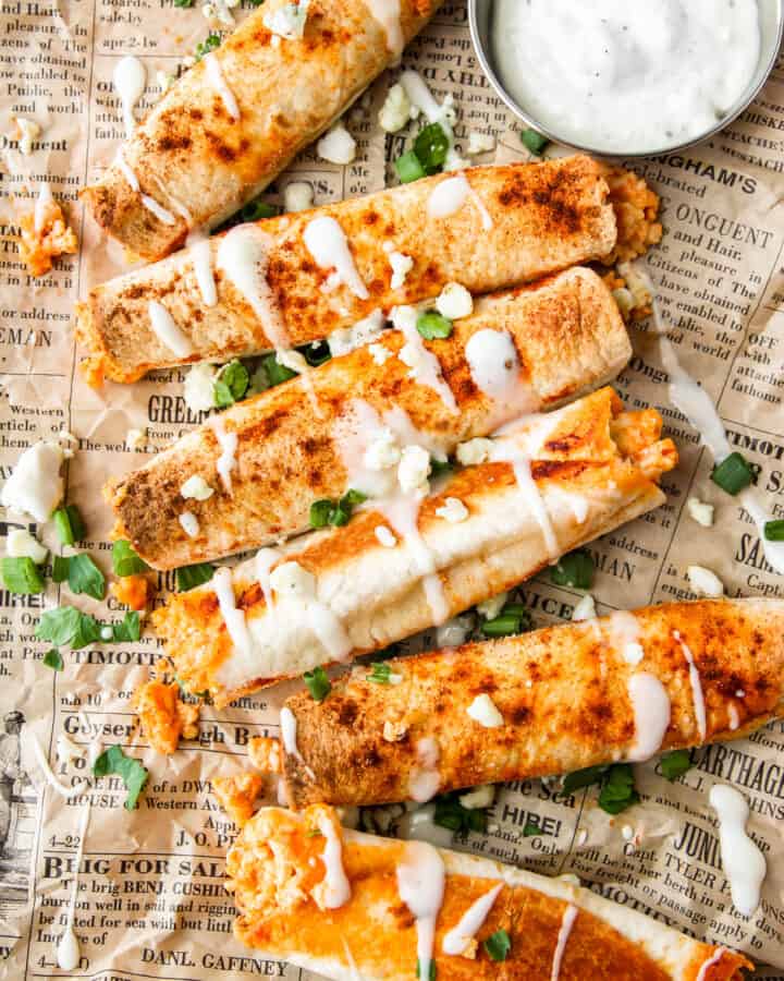 overhead shot of buffalo chicken taquitos with ranch drizzled and crumbled blue cheese, scallions, and parsley on brown newsprint paper.