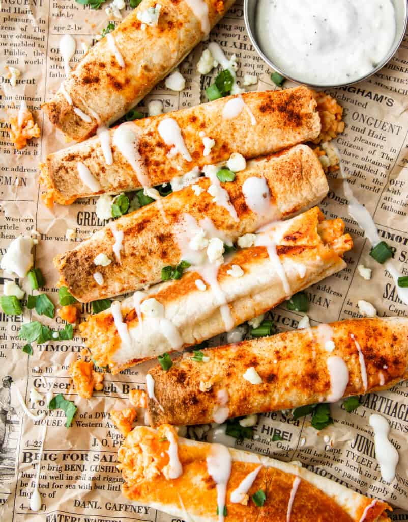 overhead shot of buffalo chicken taquitos with ranch drizzled and crumbled blue cheese, scallions, and parsley on brown newsprint paper.