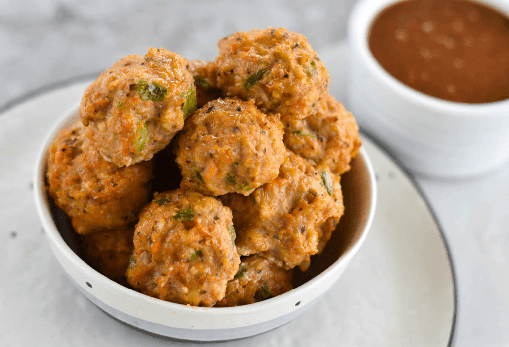 a pile of the meatballs in a white bowl with the dipping sauce in the back.