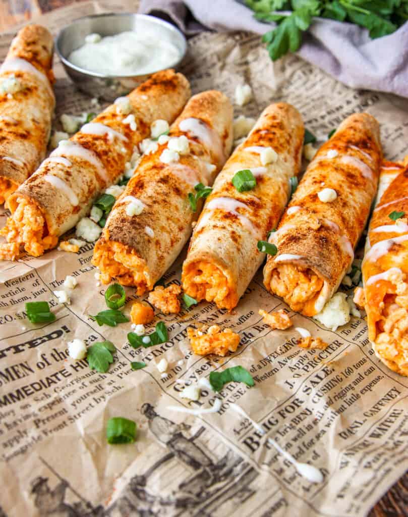 angled shot of buffalo chicken taquitos with ranch drizzled and crumbled blue cheese, scallions, and parsley on brown newsprint paper.