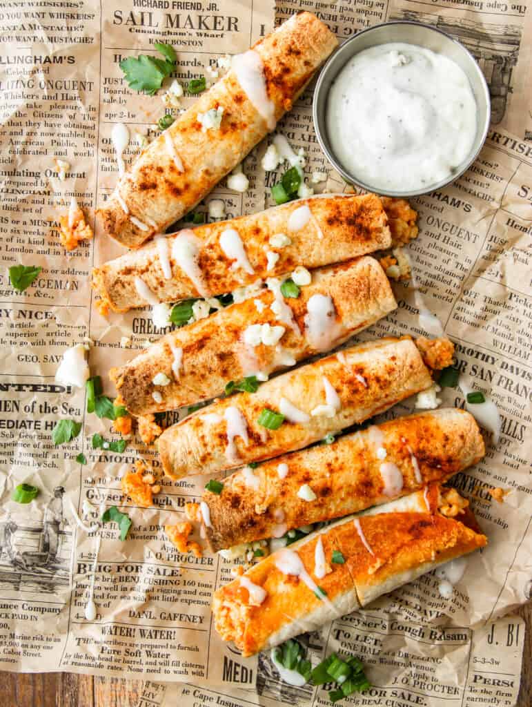 overhead shot of buffalo chicken taquitos with ranch drizzled and crumbled blue cheese, scallions, and parsley on brown newsprint paper. A small bowl of blue cheese dip.