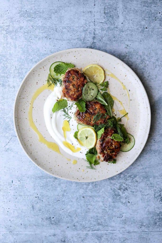 overhead shot of the chicken koftas on a speckled plate with the yogurt sauce, herbs and lemons scattered on top.