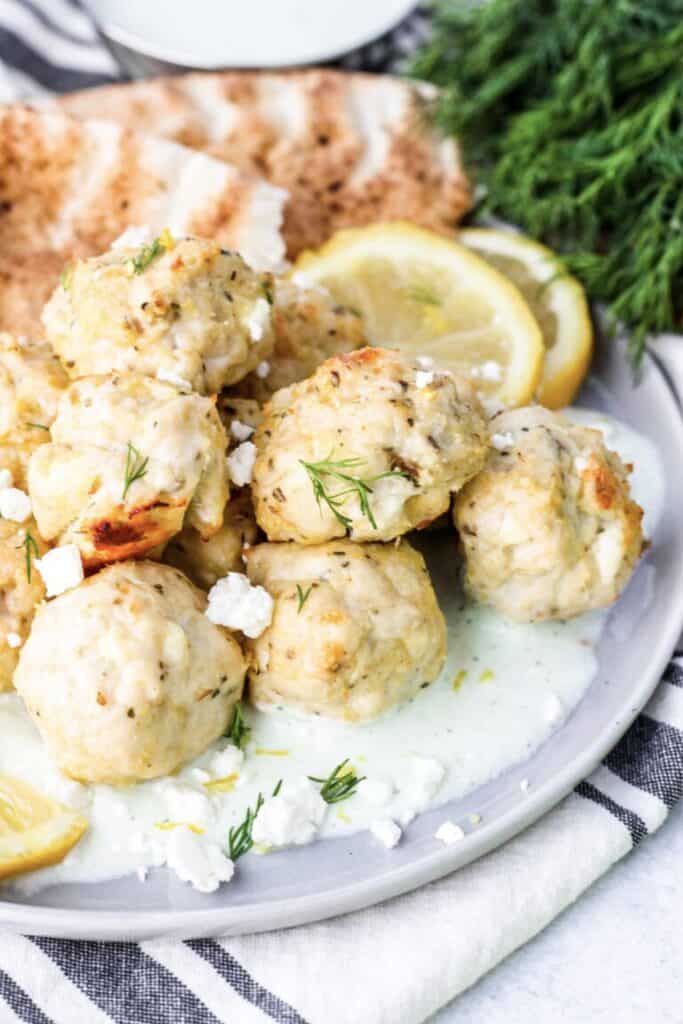 Greek chicken meatballs on a plate with cucumber sauce, a grey and white linen napkin, dill, lemon and pita in the back.
