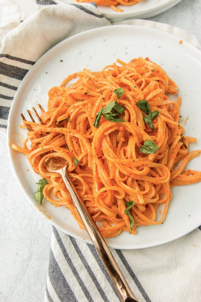 overhead shot of the creamy roasted red pepper sauce over linguini on a small white plate with a copper fork. One a blue and white stripped napkin.