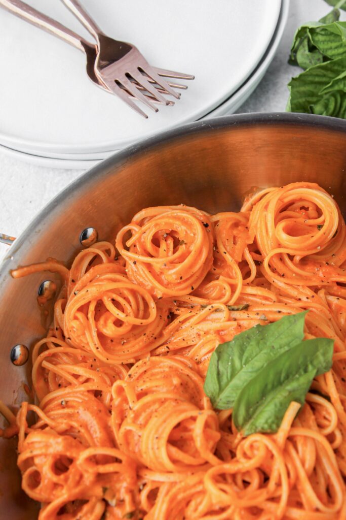 overhead shot of spiraled linguine in the creamy roasted red pepper sauce in a stainless steel pan. Forks and plates off to the side.