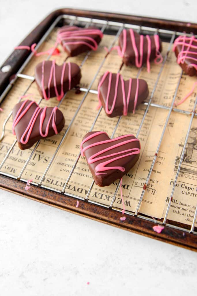 heart shaped cookie dough bites covered in chocolate and a pink drizzle. On a baking rack on a cookie sheet with brown newsprint paper underneath.