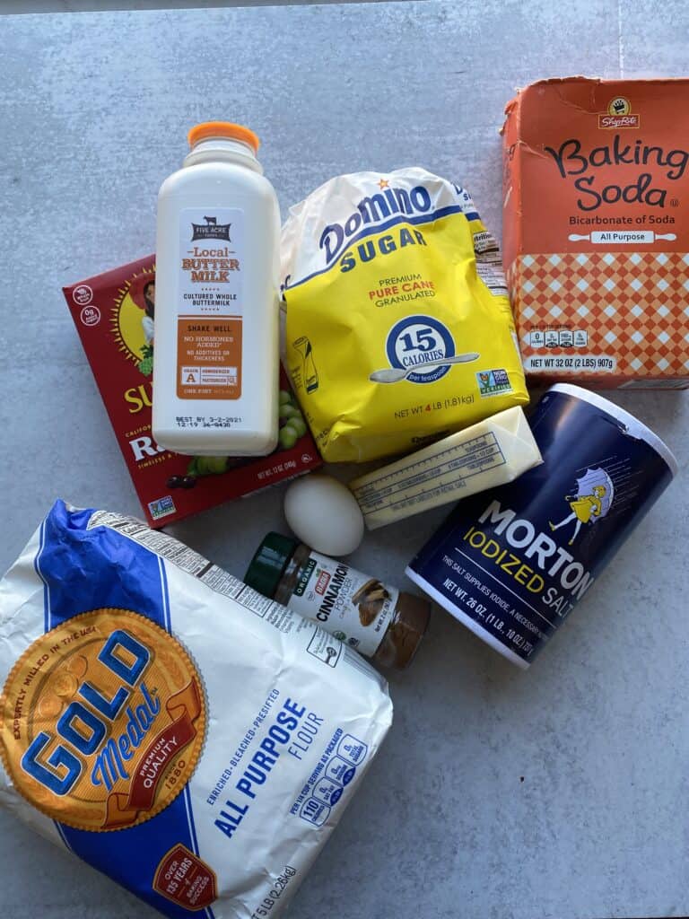 overhead shot of ingredients. Buttermilk, raisins, flour, cinnamon, an egg, butter, sugar, baking soda, salt.