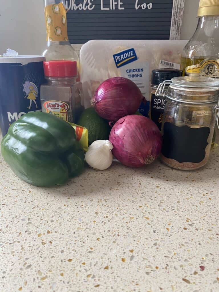 Ingredients on the counter. Salt, green pepper, oregano, packet of chicken bouillon, sour orange mix, one lime, head of garlic, 2 red onions, pack of chicken thighs, cumin, jar of sazon, olive oil