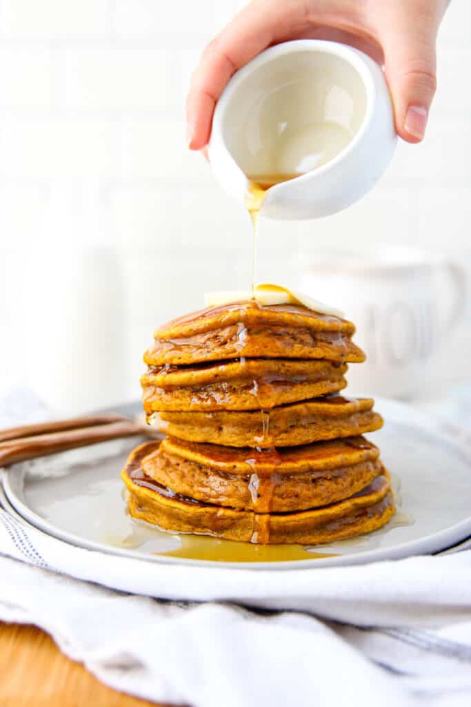 stack of 5 pumpkin pancakes on a grey round plate with maple syrup being drizzled overtop.