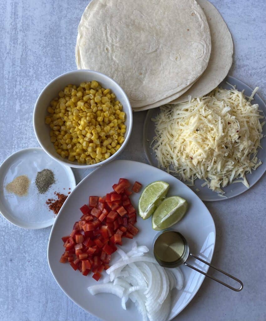 overhead shot of the ingredients, corn, seasonings on a white plate, flour tortillas, pepper jack, red peppers, onions, limes, and olive oil.