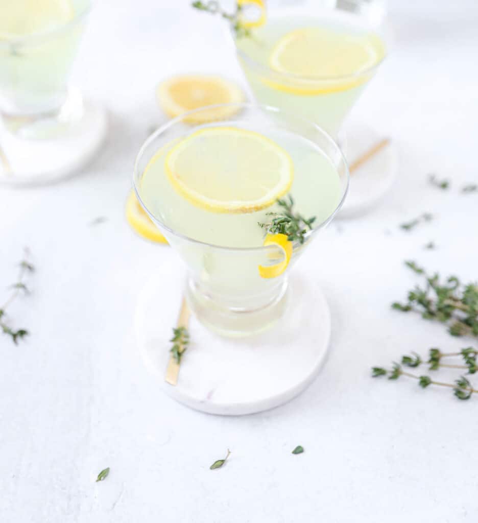 limoncello martini on a white coaster, with two martinis in the background and fresh thyme scattered. 