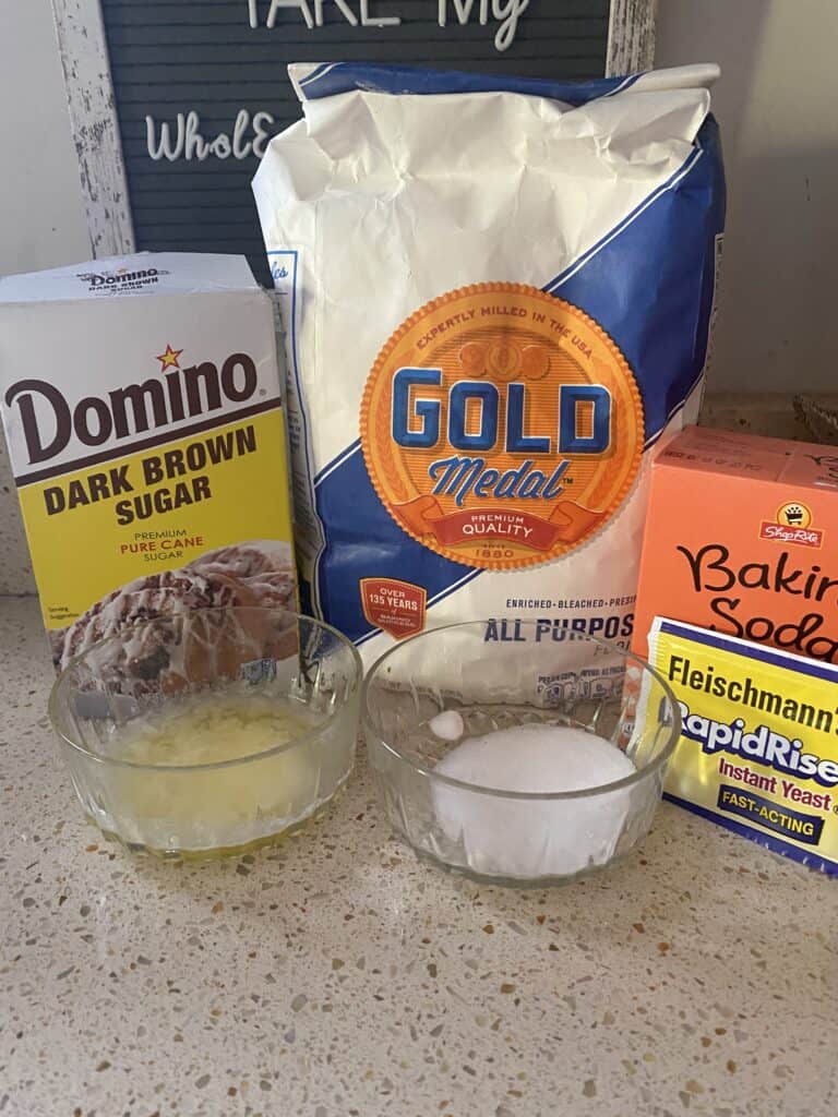 photo showing the ingredients on the counter: box of brown sugar,  melted butter, salt, bag of flour, box of baking soda, packet of rapid rise yeast.