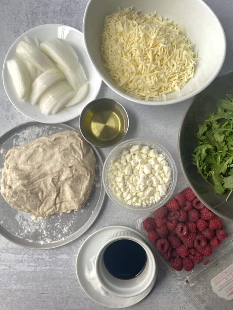 overhead shot of the pizza ingredients: slice white onions, Mozzarella cheese, olive oil, goat cheese, pizza dough, arugula, raspberries, sugar, and balsamic vinegar. 