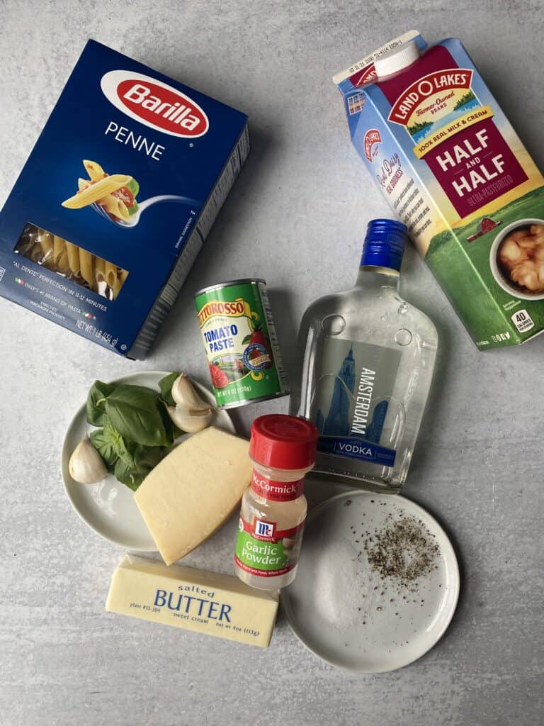 overhead shot of the ingredients on a grey surface. Box of penne, tomato paste, Parmesan, basil leaves, garlic cloves, butter, garlic powder, salt, pepper, vodka, half & hald