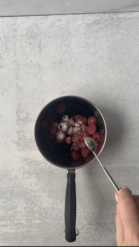 overhead shot of a small pot with the balsamic vinegar, raspberries, and pouring in the teaspoon on sugar.