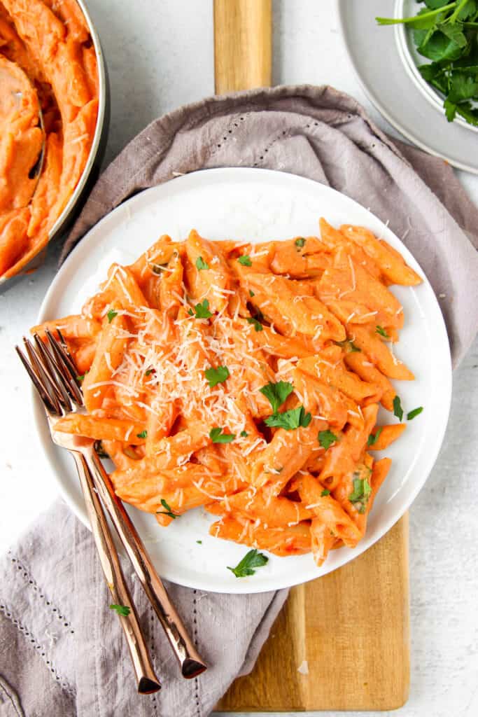 overhead shot of round white plate with the pasta, two rose gold forks, a grey napkin, on a wood cutting board. In the upper corners some parsley and the edge of the pan.