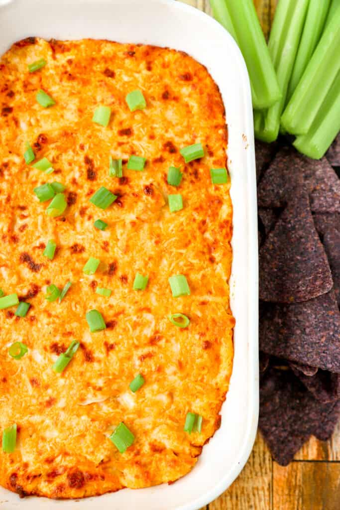 buffalo chicken dip in a white casserole dish with blue corn tortilla chips and celery.