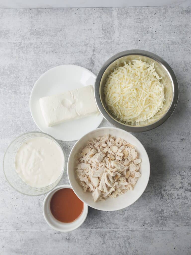 ingredients on a cement surface: cream cheese, Mozzarella cheese, ranch dressing, hot sauce, shredded rotisserie chicken.