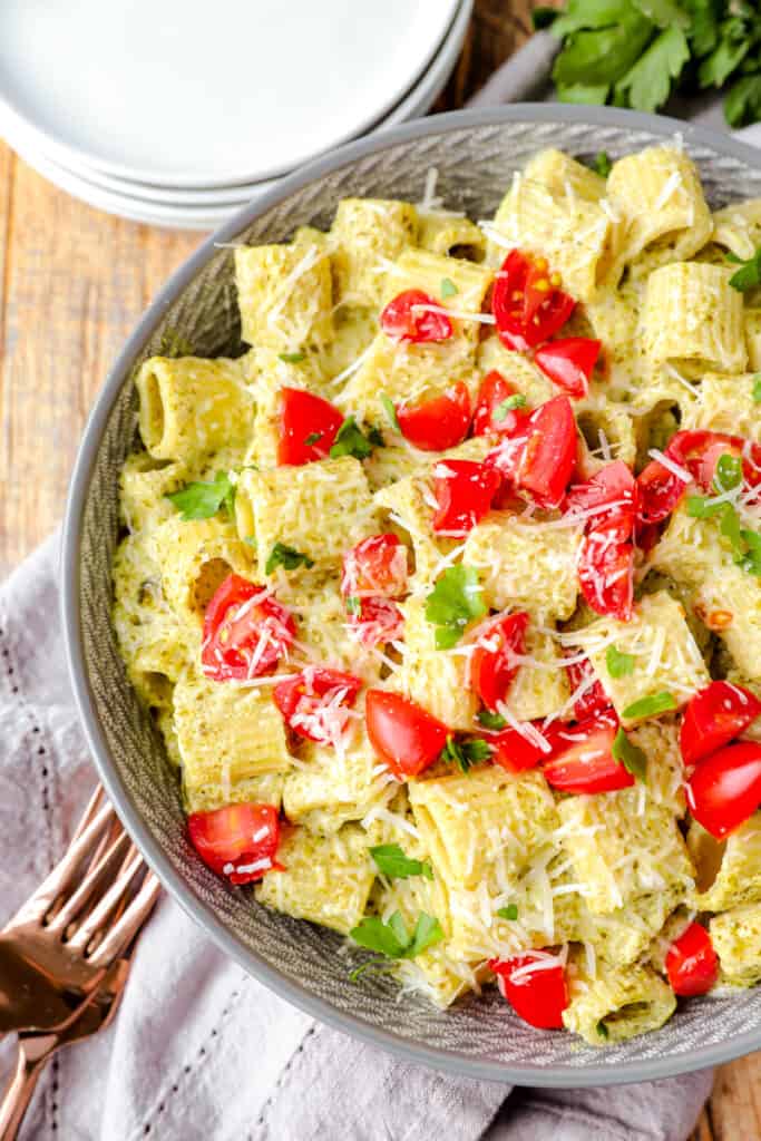 overhead shot of creamy pesto pasta in a grey bowl with diced tomatoes on top on a wood surface